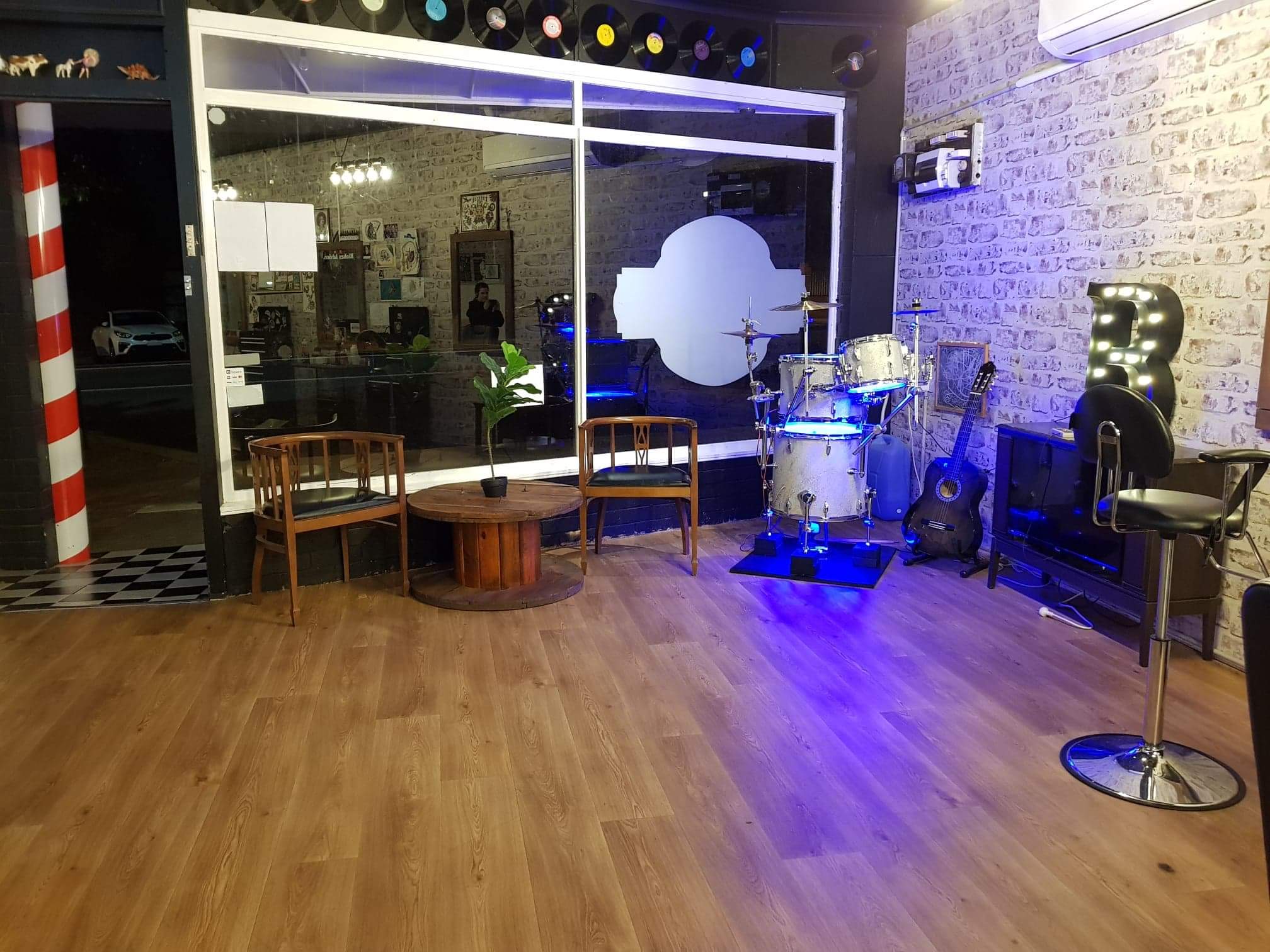 The Brother Fox waiting area at night: barber pole, vintage chairs, a lit drum kit, a guitar and a marquee letter B against white brick