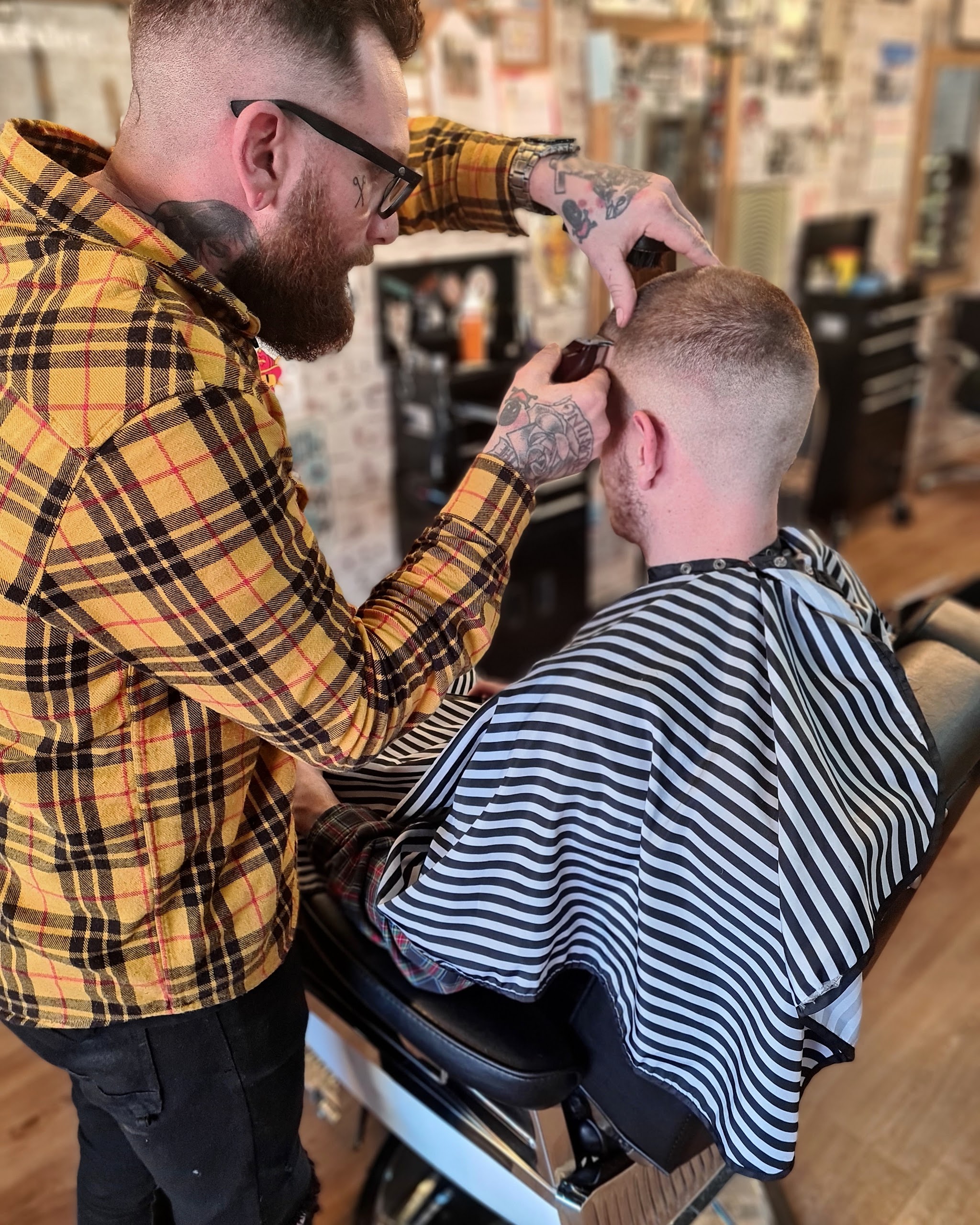 A tattooed Brother Fox barber clipper-cutting a skin fade on a client in a striped cape